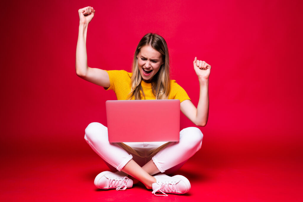 Happy young woman with winning gesture sitting on the floor with crossed legs and using laptop on red background.